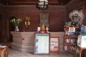 Das Bild zeigt einen Raum mit Idolen, Textfeldern und einem Rahmen an einer Wand, Blumenvasen mit Blumen, einen Wasserfilter auf einem Hocker, einen Tisch mit einem Buch, Holzregale voller Bücher, ein Fenster, eine Decke mit einer Lampe, einen Boden und eine Theke.