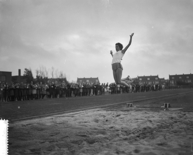Eine Frau in der Luft, die über eine Weitsprung-Anlage auf einer Bahn springt, umgeben von einer Gruppe von Menschen auf dem Boden und Gebäuden, Bäumen und einem bewölkten Himmel.