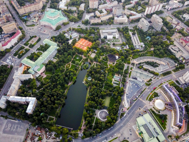 Luftaufnahme einer Stadt mit dichtem Baumbestand, Straßen, Fahrzeugen und Grünflächen, die sich um einen Park mit üppigen Bäumen und Gras gruppieren.