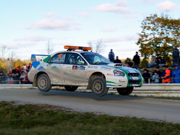 Ein weißes, grünes und blaues Rallye-Auto mit Aufschrift fährt eine Schotterstraße entlang, umgeben von Gras, mit einer Menschenmenge im Hintergrund und einem Geländer, unter einem Himmel mit Bäumen und Wolken.