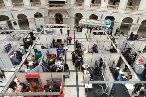 Eine große Gruppe von Menschen auf einer internationalen Fachmesse in einem Raum mit Ständen, Bannern, Tischen, Stühlen, Luftballons und einem Gebäude mit Bögen, Säulen, Geländern und einem Bildschirm im Hintergrund.