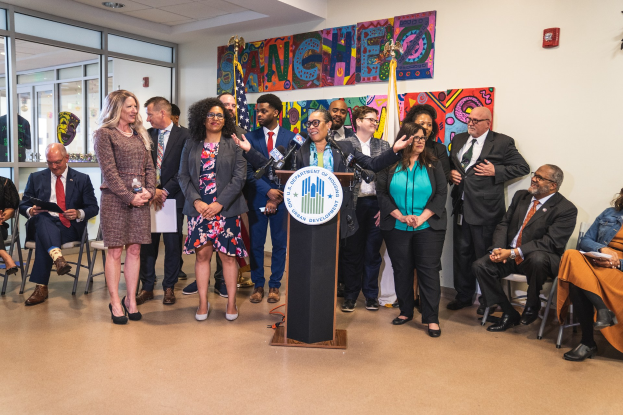 Gruppe von Menschen in einem Raum mit einem Podium, einem Mikrofon und einer Tafel mit der Aufschrift "Sancho Stadtrat tritt dem Stadtrat bei", umgeben von Flaggen, Stühlen und einem Feuerlöscher.