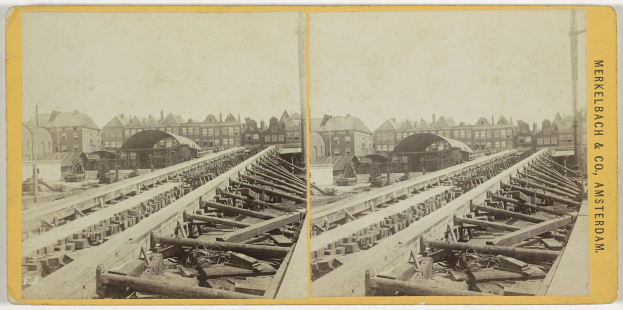 Schwarz-weiß-Foto einer Zugstrecke mit Gebäuden im Hintergrund, Text 'Merkelbach & Co, Co. Amsterdam' auf der rechten Seite und verstreuten hölzernen Gegenständen im Vordergrund.