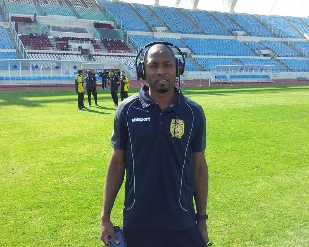 Ein Mann in einem Headset, wahrscheinlich der Trainer der ghanaischen Nationalmannschaft, steht auf einem Fussballfeld in der Nähe eines Stadions und hält ein Objekt, whährend im Hintergrund Menschen zu sehen sind und der Himmel sichtbar ist.