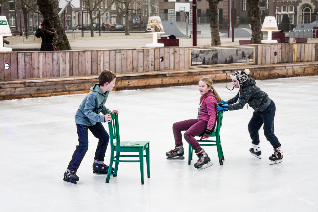 Kinder beim Skifahren im Vordergrund eines Spielplatzes, mit drei Kindern und zwei Stühlen in der Mitte und Gebäuden, Bäumen, Bänken, Pfosten und einem Basketballfeld im Hintergrund.