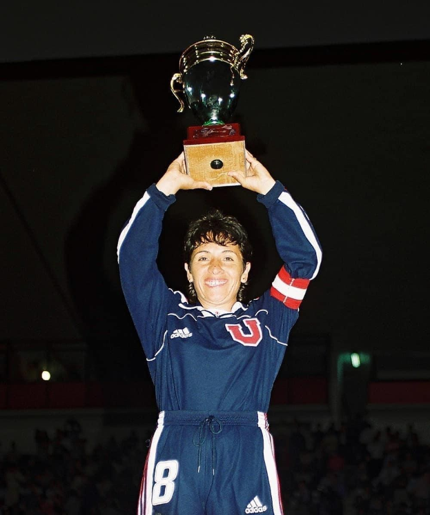 Eine Frau in einem blauen Kleid hält eine Trophäe hoch und lächelt, umgeben von einer jubelnden Menge in einem hell erleuchteten Stadion.
