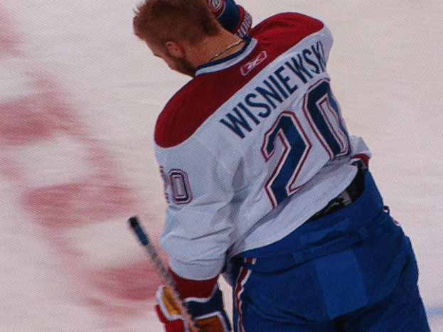 Ein Eishockeyspieler in einem "Wisniewski"-Trikot steht auf dem Eis und hält einen Hockeyschläger, mit einem konzentrierten und entschlossenen Gesichtsausdruck.