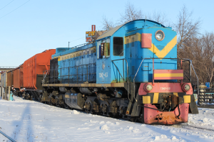 Ein blauer und gelber Zug fährt auf schneebedeckten Schienen mit einem Geländer auf der rechten Seite, Bäumen im Hintergrund, einer fernen Brücke und einem klaren Himmel darüber.