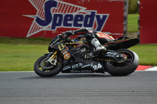 Eine Person in Helm und Schutzkleidung fährt ein Motorrad auf einer Rennstrecke, mit Gras und einer Tafel mit Text im Hintergrund.
