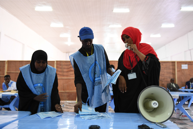 Menschen um einen Tisch mit Megafon und Papieren an einer Wahllokal, mit anderen sitzend und stehend im Hintergrund unter Deckenleuchten.