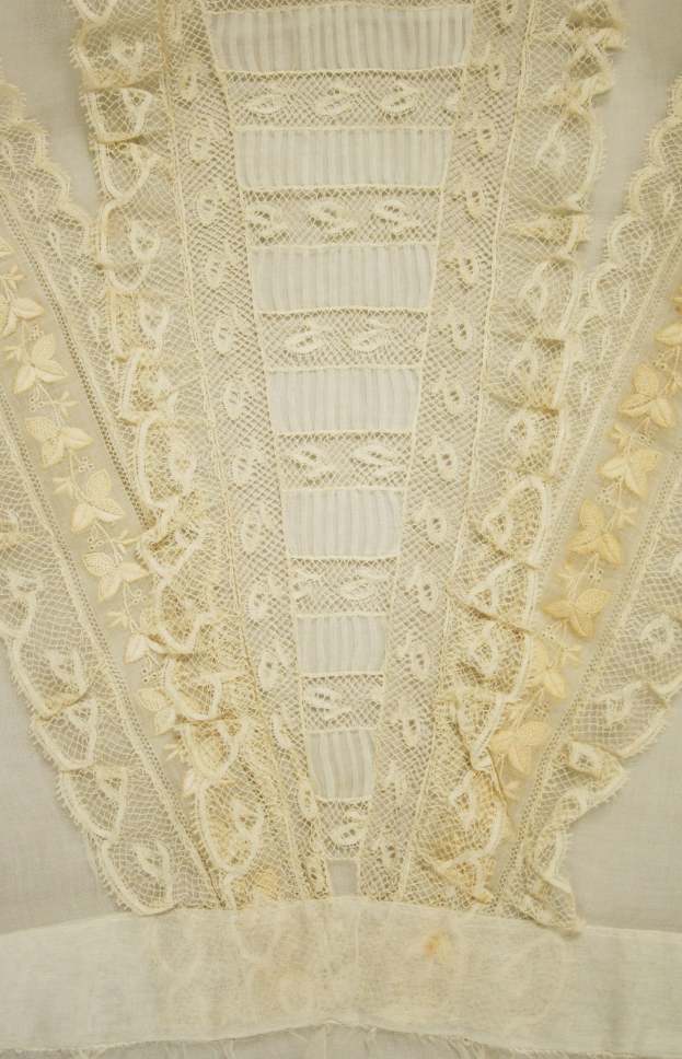 Nahaufnahme eines weißen Kleides mit zarten, detailreichen Spitzenverzierungen auf einem leichten, luftigen Stoff.