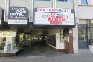 Das Lincoln Theater in Berlin, Deutschland, ein Gebäude mit Glasfenstern und -türen sowie einer Tafel mit Text, die verschiedene Objekte zeigt und den Eindruck einer pulsierenden Stadtlandschaft vermittelt.
