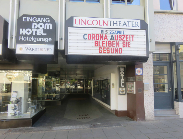 Das Lincoln Theater in Berlin, Deutschland, ein Gebäude mit Glasfenstern und -türen sowie einer Tafel mit Text, die verschiedene Objekte zeigt und den Eindruck einer pulsierenden Stadtlandschaft vermittelt.