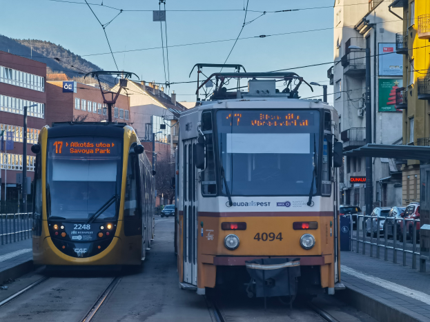 Zwei gelbe und weiße Straßenbahnen auf einer Stadtstraße mit hohen Gebäuden, Fahrzeugen, Geländern, Pfählen, Schildern, Bäumen, einem Hügel und dem Himmel im Hintergrund.
