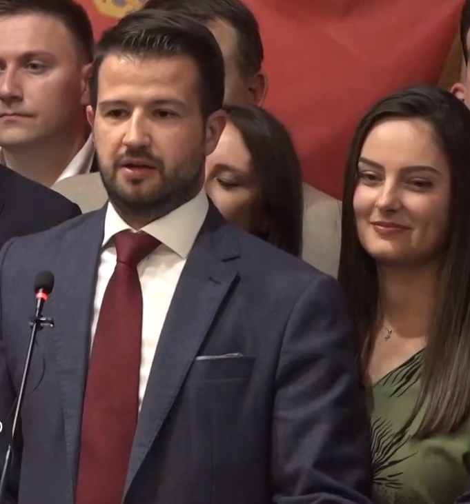 Ein Mann im Anzug spricht in ein Mikrofon an einem Podium, mit einer Menge und einem Banner im Hintergrund.