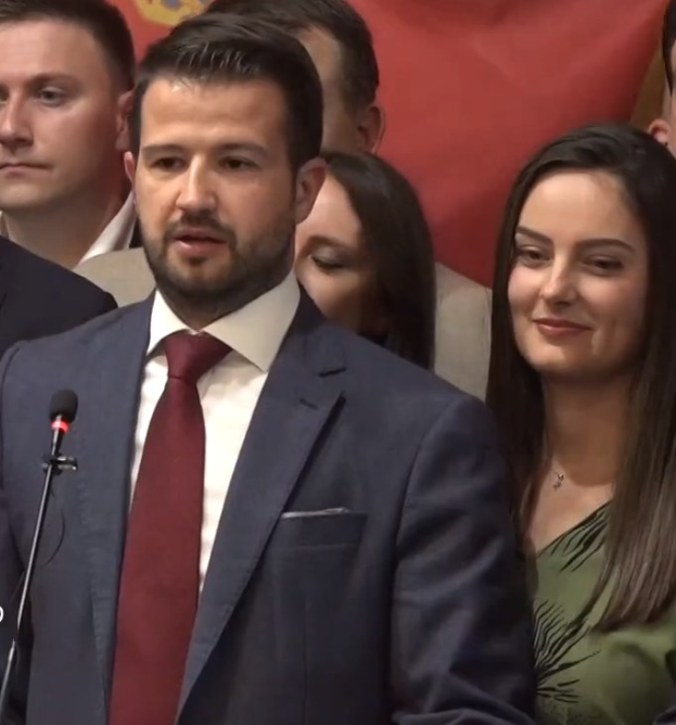 Ein Mann im Anzug spricht in ein Mikrofon an einem Podium, mit einer Menge und einem Banner im Hintergrund.