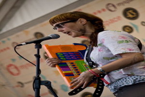 Eine Frau in einem weißen Hemd und Sonnenbrille singt in ein Mikrofon, während sie ein Piano spielt, mit einem Plakat an der Wand hinter ihr.