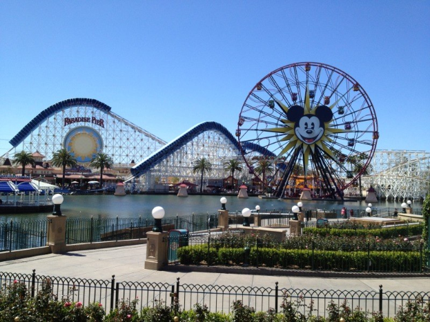 Der Disneyland California Adventure Vergnügungspark mit einem Riesenrad, Achterbahn, Grünflächen, Laternenmasten, einem Gewässer und einem strahlend blauen Himmel.