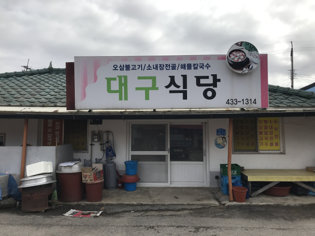 Kleines Gebäude mit koreanischem Restaurant-Schild, Tür, Fenstern und nahen Containern, Eimern, Tisch und Drähten unter einem bewölkten Himmel.