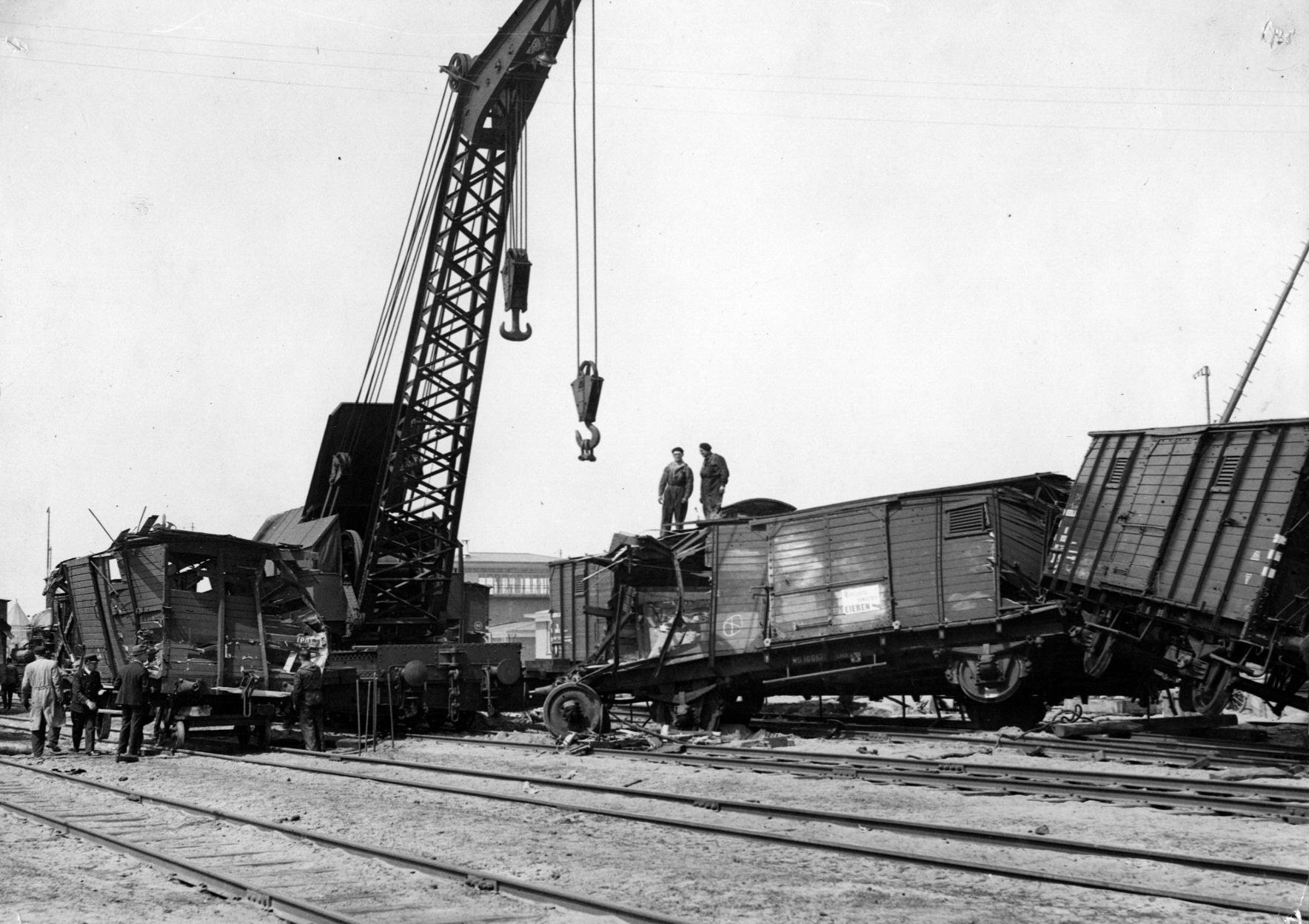 Schwarzes und weißes Bild einer Zugkatastrophe mit einem Kran, der einen Zugwagen auf die Schienen hebt, mehreren Menschen in der Nähe und im Hintergrund Gebäude und einen klaren Himmel.
