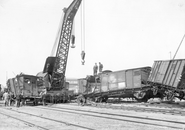 Schwarzes und weißes Bild einer Zugkatastrophe mit einem Kran, der einen Zugwagen auf die Schienen hebt, mehreren Menschen in der Nähe und im Hintergrund Gebäude und einen klaren Himmel.