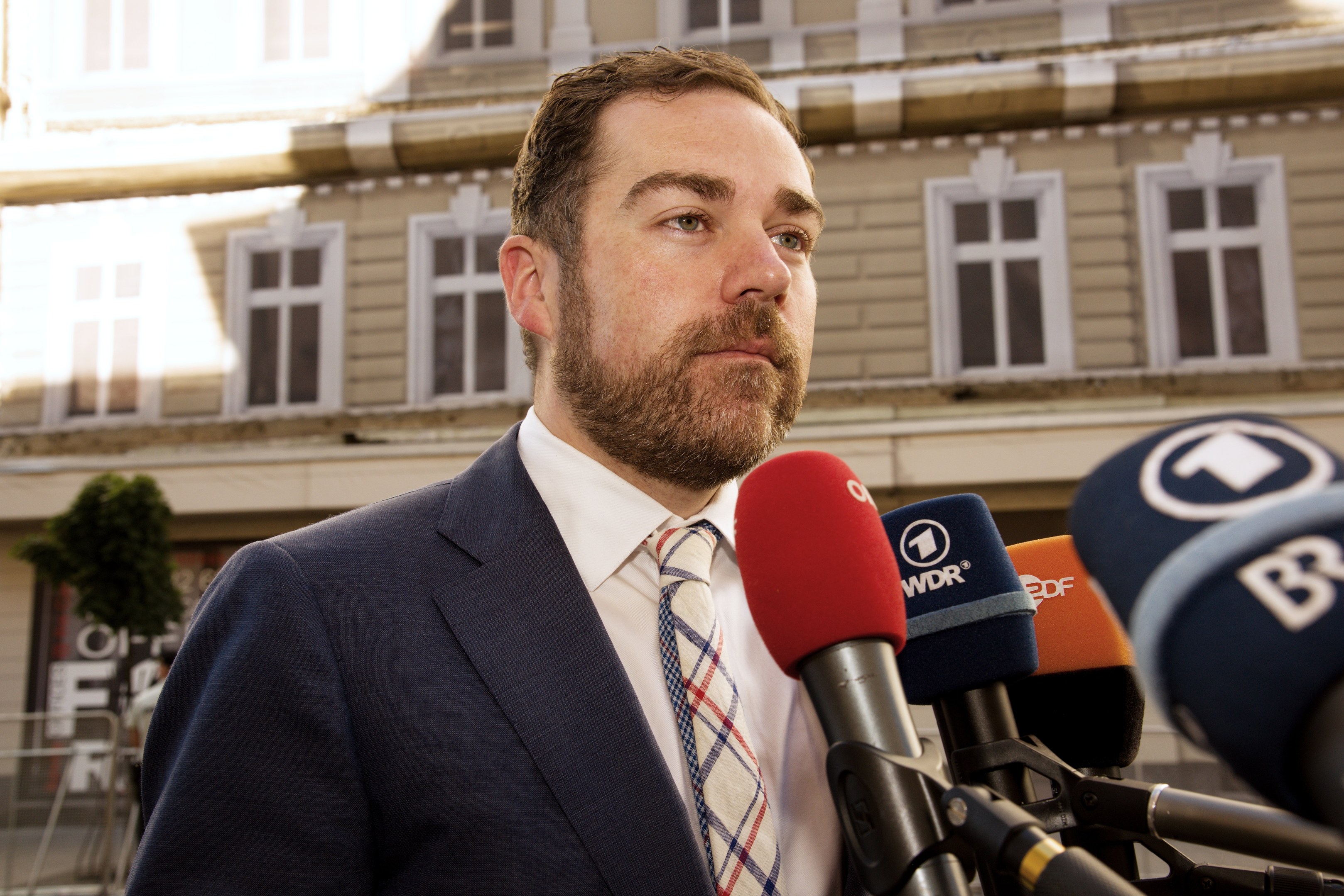 Ein Mann in Anzug und Krawatte spricht vor einem Gebäude zu den Medien, wahrscheinlich wegen einer dreijährigen Haftstrafe.