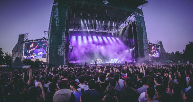 Eine große Menschenmenge schaut bei einem Musikfestival auf einer hell erleuchteten Bühne mit Instrumenten und Bildschirmen, die vor einem Gebäude, Bäumen und einem blauen Himmel aufgebaut sind.