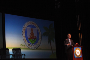 Ein Mann in weißem Hemd und schwarzer Jacke steht an einem Rednerpult, möglicherweise sprechend, mit Stühlen im Vordergrund und einem schwarzen Hintergrund, der ein Board oder ein Projektionsschirm sein könnte, was eine Konzerthalle vermuten lässt.