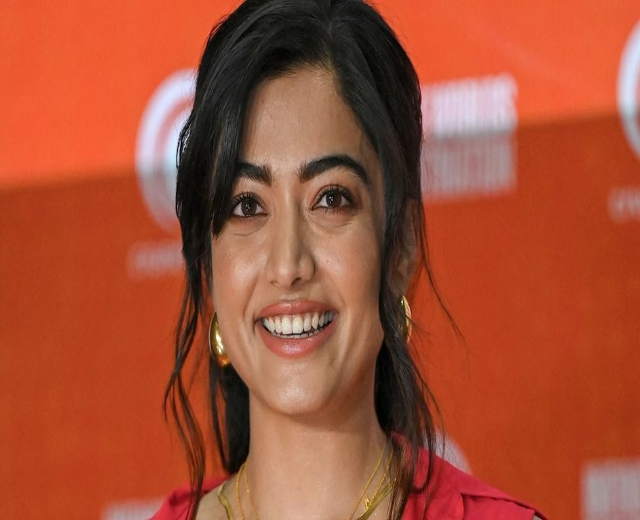 Eine Frau in einem roten Kleid mit einem eleganten Dutt, schwarzen High Heels und natürlichem Make-up lächelt in die Kamera vor einem gelben Banner mit der Aufschrift "Sonam Kapoors beste Schönheits Looks vom Cannes Film Festival."