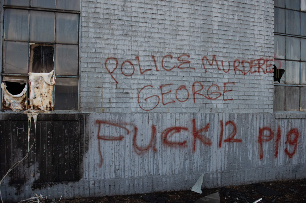 Ein Gebäude mit Graffiti, das "Polizei ermordet George Fuck 12 P19" auf der Seite trägt, mit Fenstern und verschiedenen Gegenständen vorne drauf.