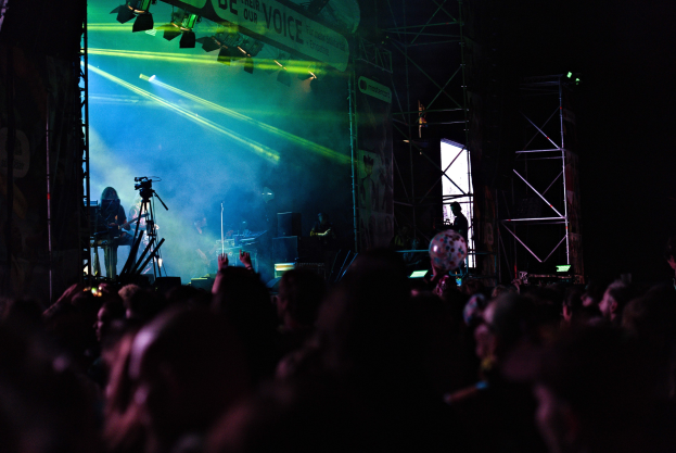 Eine Menge schaut Musikern auf einer hell erleuchteten Bühne bei einem Konzert zu, mit Musikinstrumenten, Mikrofonen und einem Banner.