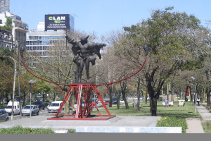 Eine Skulptur mit Metallverzierung in einer städtischen Umgebung, die Bäume, Gebäude mit Fenstern, Fahrzeuge auf der Straße, Bänke, Menschen in Kleidung, Gras, Pfosten und einen Himmel zeigt, mit einem Plakat mit Text im Vordergrund.