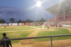 Baseballspiel in einem Stadion mit Zuschauern auf den RÀngen, BÀumen, Masten, Lichtern, Werbetafeln und einem klaren blauen Himmel im Hintergrund.