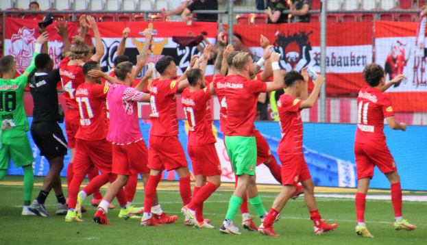 Gruppe von Fußballspielern, die ein Tor feiern, auf einem Feld, einige halten Flaschen oder klatschen, mit Bannern, einem Zaun, Stühlen und Deckenleuchten im Hintergrund.