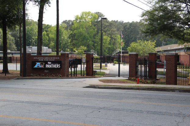 Der Eingang zu einer Schule mit einem Schild an der Seite, umgeben von Bäumen, Straßenmasten, Straßenlaternen, Gittern, Namensschildern, Kraftfahrzeugen auf der Straße, Verkehrskegeln, Pflanzen, Gras, Gebäuden und einem klaren blauen Himmel.