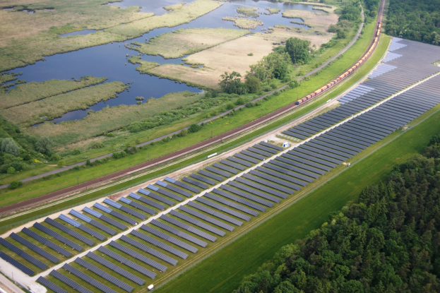 Luftaufnahme einer Solar-Farm mit Panelen, umgeben von Bäumen, Gras, Wasser und einer nahen Bahnschiene.