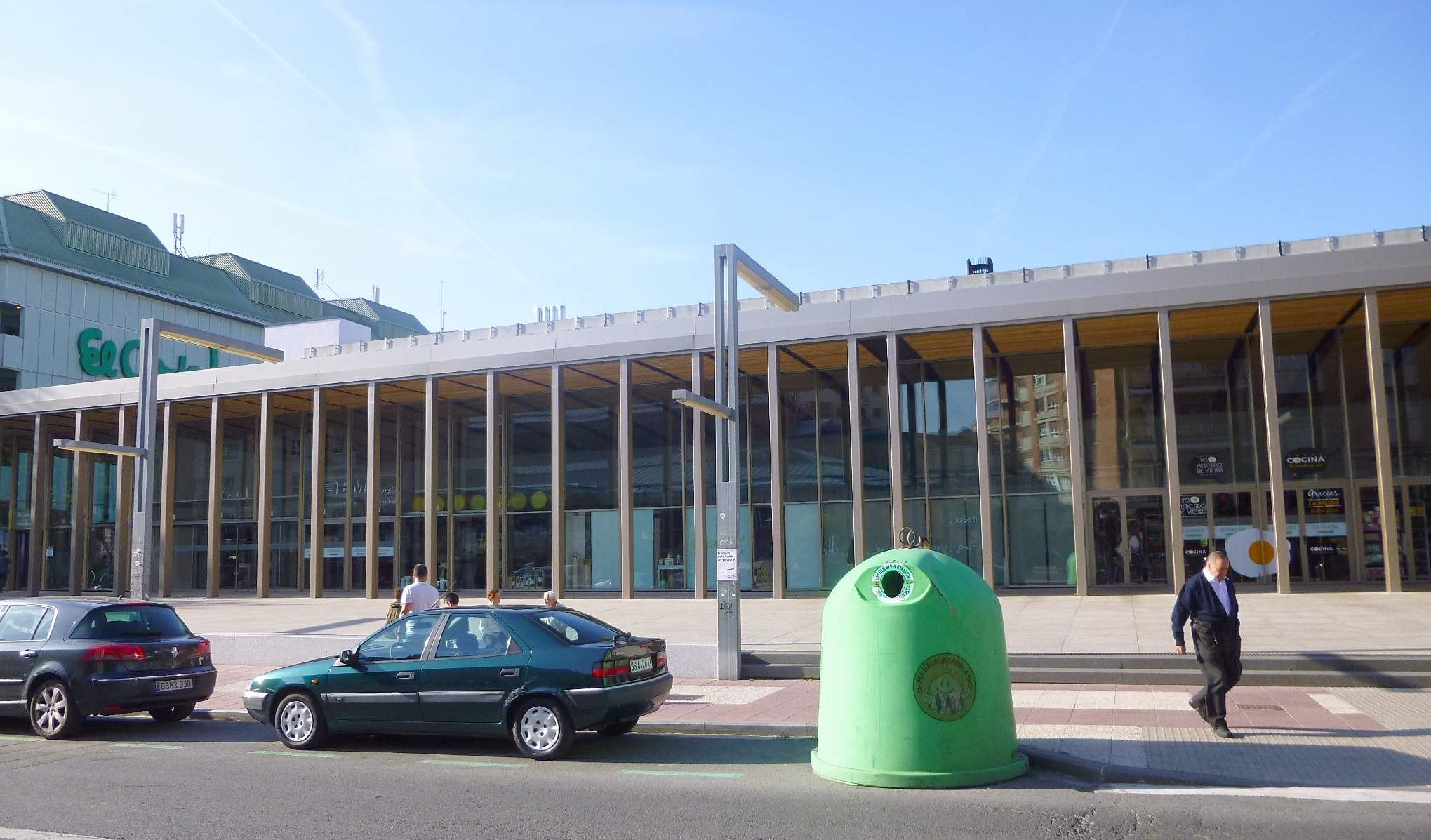 Grüner Müllcontainer auf einer Stadtstraße neben einem glaswandigen Gebäude mit Säulen, mit Fahrzeugen und Fußgängern im Vordergrund und einem bewölkten Himmel im Hintergrund.