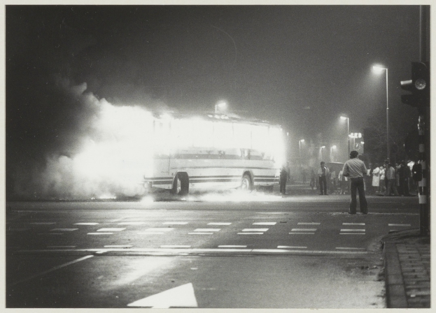 Schwarz-weiß-Foto eines brennenden Busses am Straßenrand mit einer Gruppe von Menschen auf dem Gehweg rechts und beleuchteten Straßenlaternen im Hintergrund.