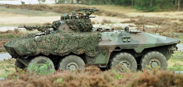 Militärisches Personalfahrzeug mit einer montierten großen Waffe, das auf einer Straße neben einem Wald fährt.