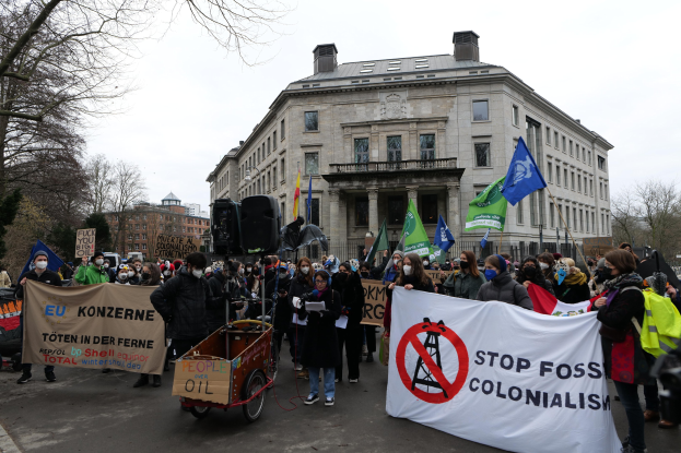 Große Gruppe von Menschen bei einer Demonstration gegen fossile Brennstoffe, die Schilder und Fahnen halten, mit einem Fahrzeug im Vordergrund und Gebäuden, Bäumen und einem klaren blauen Himmel im Hintergrund.