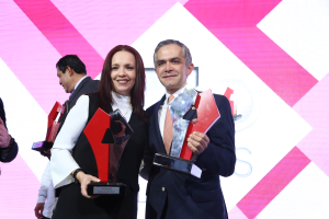 Ein Mann und eine Frau stehen nebeneinander auf einer Bühne, jeder hält eine Trophäe und lächelt, mit einem Bildschirm im Hintergrund.