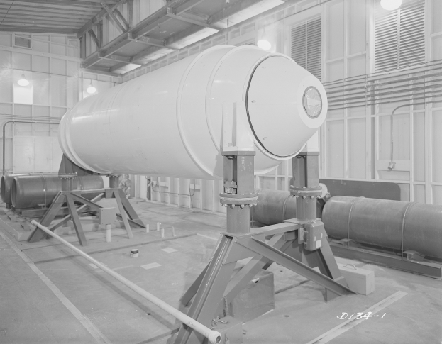 Eine große zylindrische Wasserstoffbombe auf einem Ständer in einer industriellen Fabrikumgebung, mit sichtbaren Rohren, Lampen und einer Wand im Hintergrund und Text unten.