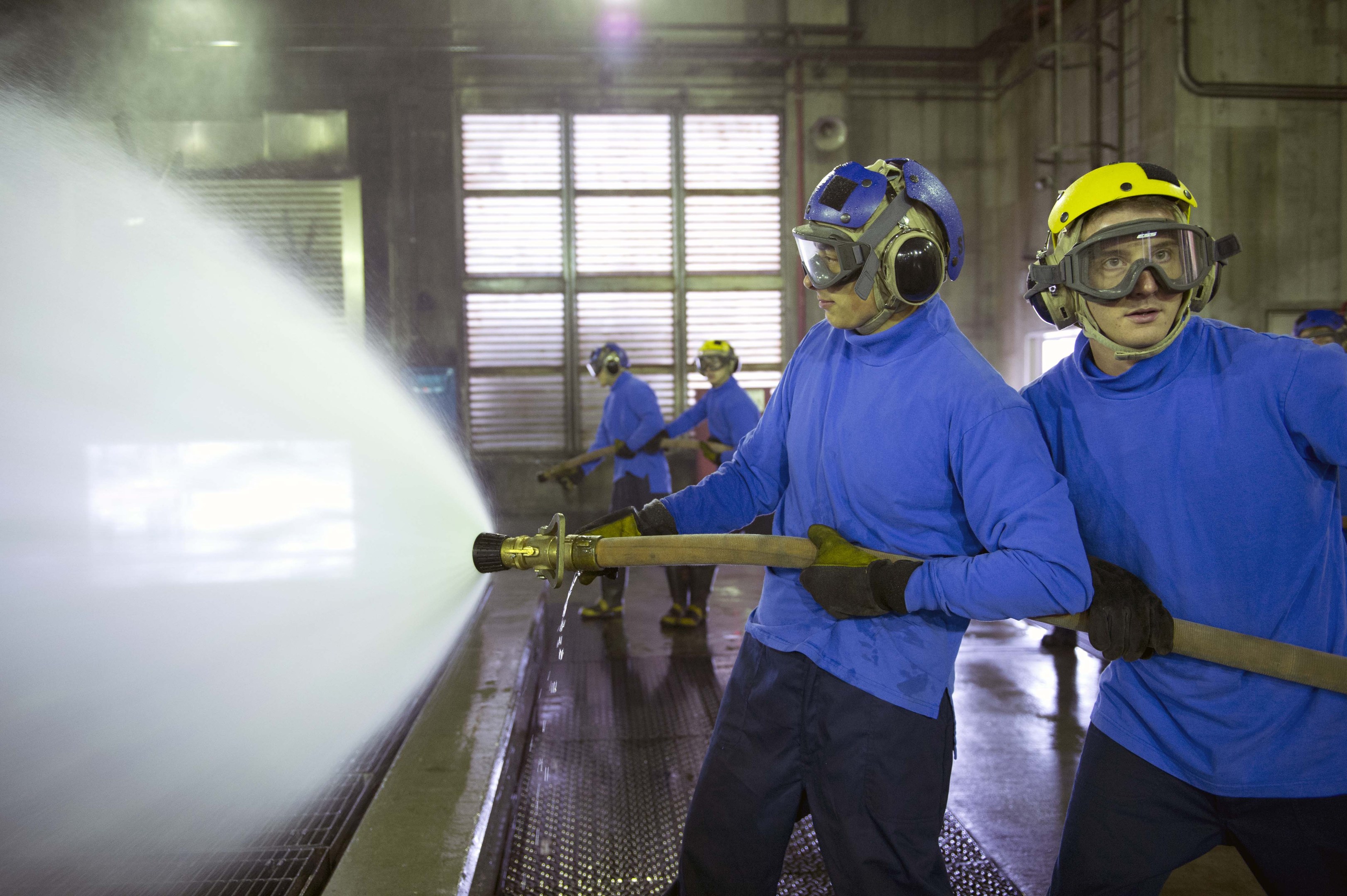 Gruppe von Männern in blauen Hemden und gelben Helmen, die an einer Maschine arbeiten, einer sprüht Wasser auf den Boden in einer Fabrik mit sichtbaren Wänden, Fenstern, Rohren und Lampen.