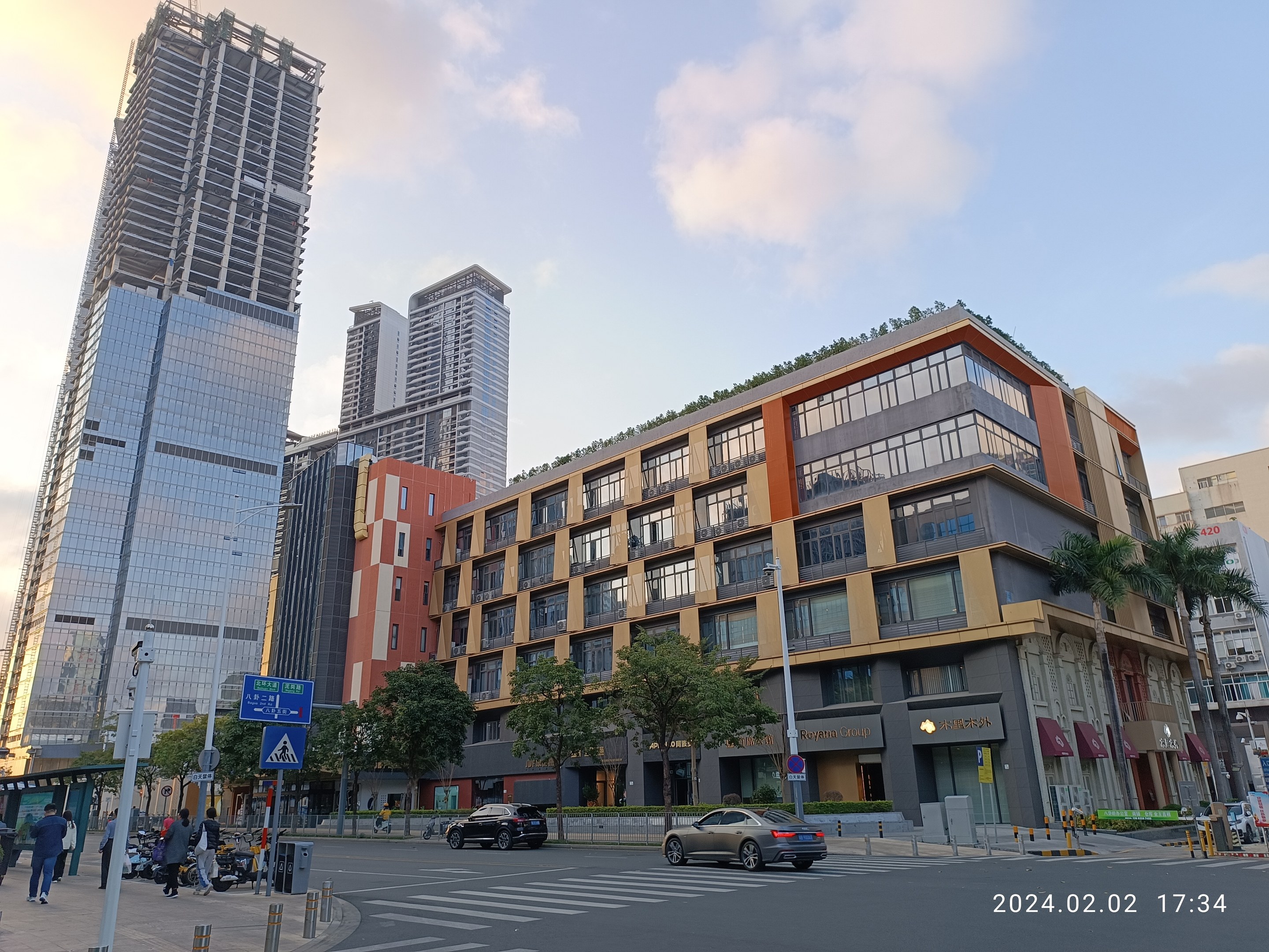 Eine belebte Stadtstraße mit hohen Gebäuden im Hintergrund, Fahrzeuge auf der Straße, Fußgänger auf dem Gehweg und Bäume, die die Straße säumen, unter einem bewölkten Himmel, mit einem Bürogebäude mit grüner Dachfläche im Vordergrund.