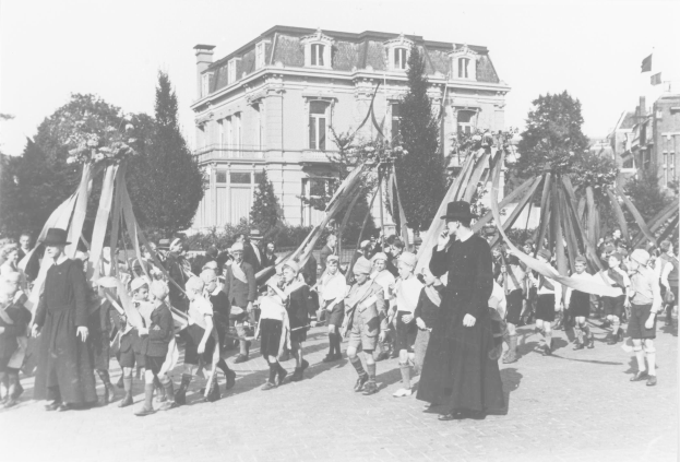 Schwarzes und weißes Bild einer Gruppe von Menschen, die eine Straße entlanggehen und Fahnen tragen, mit Hüten, Bäumen, Gebäuden und einem Maibaum im Hintergrund.