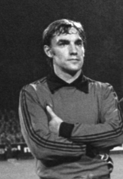 Schwarzes und weißes Foto eines Mannes, der mit verschränkten Armen auf einem Fußballfeld steht und von einer Gruppe sitzender Menschen im Hintergrund umgeben ist.