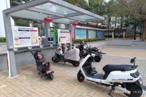 Mehrere Elektro-Scooter vor einer Bushaltestelle geparkt, umgeben von Bäumen, Gebäuden und einem klaren blauen Himmel, mit Schildern in der Nähe.