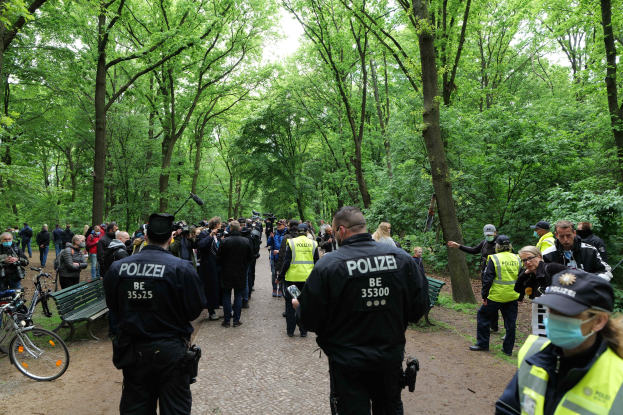 Polizisten vor einer Menge während einer Outdoor-Veranstaltung, mit Fahrrädern und einer Bank im Vordergrund, Bäumen und Himmel im Hintergrund.