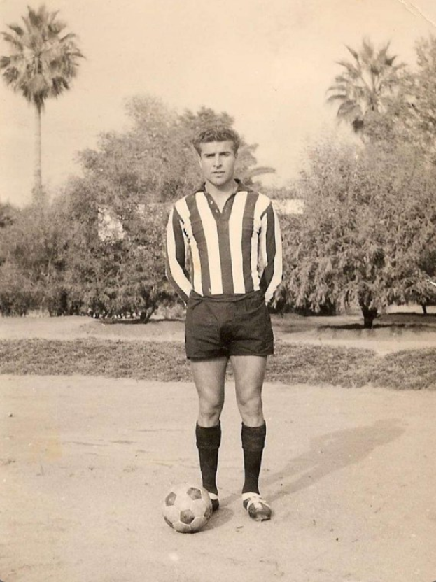 Schwarzes und weißes Foto eines Mannes, der auf Gras steht und einen Füüball hölt, mit Bäumen und Himmel im Hintergrund.