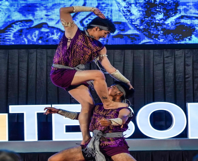 Ein Mann und eine Frau in lila Kleidern führen Akrobatik auf der Bühne vor, wobei der Mann die Frau in seinen Armen hält, während eine Menge von unten hinter einem schwarzen Vorhang und einem Bildschirm zusieht.